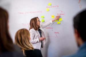 Carola Öberg, tidigare projektledare på Science Park Jönköping står framför en vägg med postit och texter och presenterar vad som står på väggen. På väggen står ord som "impact". "material" och "framtiden"